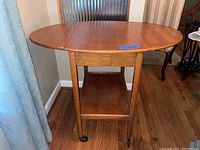 Front view of the oak drop leaf table with rounded edges and central base shelf, showing legs with wheels.