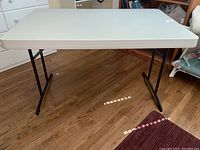 Front angled view of white folding table with black legs on wooden floor, showing table top and leg design.
