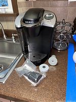 Keurig coffee maker showing side water reservoir, drip tray, control panel, with coffee maker accessories and parts on counter