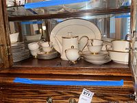 Photo of china set arranged in a wooden display cabinet including teacups with saucers, bowls, a coffee or teapot, sugar bowl, and serving platter all with gold trim.