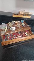 Photo showing wooden jewelry box with glass top filled with assorted men's jewelry including cuff links, pins, necklaces, pocket watch, and other small accessories