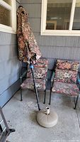 Patio umbrella closed with floral fabric beside two armchairs with matching cushions.