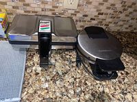 Two countertop electric appliances: rectangular Prego Pizzelle Baker and round Cuisinart waffle maker side by side on granite counter.