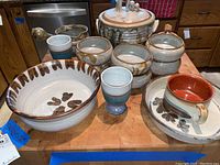 Photo showing a collection of stoneware kitchen items on a wooden surface including a large mixing bowl with floral design, a lidded casserole dish, multiple smaller bowls, a pour bowl, and two blue-glazed cups.