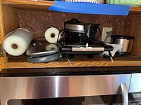 Three kitchen appliances on wooden shelf: Kitchen Gynti vacuum sealer, Oster waffle maker, and panini press.