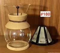 Clear glass drip coffee pot with cork insulation band around neck and base, cork trivet, and black/stainless drip cone filter shown together on wood background.