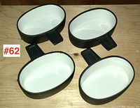 Four oval black and white enamelled cast iron dishes arranged on wooden surface showing size and shape, each with a short flat handle.