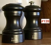 Two dark wooden Peugeot kitchen items, one pepper mill and one salt shaker, showing front view with branding visible.