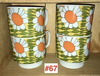 Four ceramic mugs stacked in pairs in wooden shelving, showing bold floral pattern of white petals with orange centers on yellow and green background, pedestal base visible.