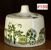 White ceramic mustard pot with lid showing green and mustard vegetable and floral pattern on one side.