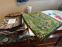 Photo of assorted fabrics and linens on wooden furniture showing varied patterns and colors including paisley and floral embroidery.