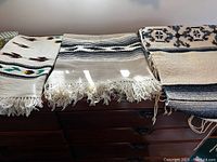 Three folded woven textiles laid out side by side on a wooden dresser showing patterns and fringes
