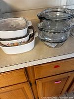 Stack of white ceramic baking dishes with clear lids, includes rectangular and square shapes, one with fruit design.