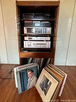 Front view of stereo cabinet displaying multiple stacked audio components with multiple vinyl records stacked in front on the floor.