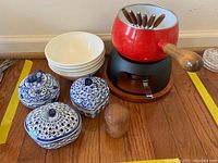 Photo of entire set showing fondue pot on black stand with 6 forks, 3 blue and white lidded dishes, white bowls stacked, and wooden pepper grinder on wooden table.