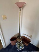 Full view of brass floor lamp with pink frosted glass shade showcasing height and design. Nearby are the basket, waste can, and flower arrangement grouped on the floor.