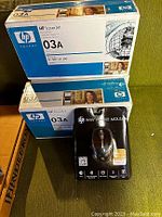 Photo showing three unopened HP 03A LaserJet Print Cartridge boxes stacked on top of each other and one black box of HP Wi-Fi Mobile Mouse on a green fabric surface.