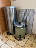 Pedal-operated stainless steel trash can, electric beverage dispenser, and marble tabletop grouped together on floor.