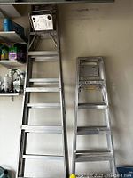 Two aluminum ladders in a garage setting, one about 6 feet tall and the other about 9 feet tall.