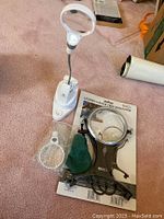 White plastic magnifier with gooseneck and lens cover alongside black magnifier in packaging and accessories on carpet