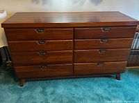 Mid-century modern wooden dresser with 8 drawers and recessed metal pull handles shown from the front on a teal carpet.