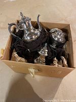 Box containing various silver serving pieces including a teapot, sugar bowl, and creamer placed on black cloths.