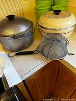 Two soup pots and two mesh strainers placed on a kitchen countertop. The background shows a yellow wall and green containers.