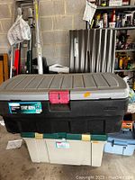 Top view showing the large black storage container with gray lid and red latch on top of a beige with green lid container.