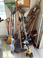 Various yard tools leaning against a wall, including rakes, shovels, broom, and tiki torches; garden tiller with small motor on the floor.