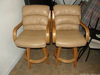 Two tan leather or faux leather padded barstools with wooden frames, rounded footrests, placed side by side on wooden floor.
