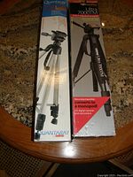 Photo showing two boxed camera tripods, Quantaray Sutro III in white box and Promaster Ultra 7000TM in black box, placed upright on a table.