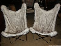 Two faux fur slingback chairs viewed from back showing light gray faux fur upholstery with darker streaks and black metal frames.