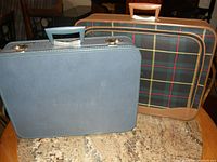 Two vintage suitcases side by side on a surface, one light blue hard shell and one plaid fabric with leather trim.