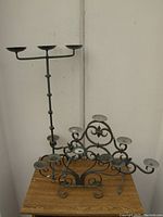 Three metal candle holders displayed on a wooden surface against a white wall, includes a small seven-cup holder, medium three-arm candelabra with curved arms, and tall segmented three-arm candelabra.