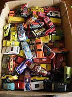 Box filled with approximately 40 loose die cast cars in various colors including yellow, red, white, purple, orange, and black. Includes race cars, trucks, and classic cars with various decals.