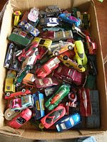 Top-down photo showing a large quantity of loose die cast cars in a cardboard box, with a variety of colors and models visible.