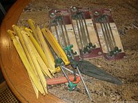 Large lot of yellow plastic and metal tent stakes with green screws, some in original packaging, others loose on table.