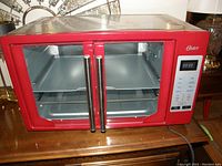 Front view of red Oster toaster oven featuring dual glass doors with stainless steel handles, digital timer, and control buttons on the right side. The interior shows two metal trays and a wire rack.