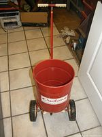 Front view of red metal Cyclone seed spreader showing cylindrical barrel, wheels, handle with white grip, and 'Cyclone' label on barrel.