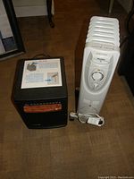 Two electric space heaters on floor - black compact heater with wood accent and white radiator heater with wheels and controls.