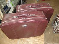 Two large maroon vintage American Tourister suitcases side by side, showing top handles, zipper closures, and rectangular shape.