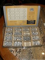 Open metal box displaying various compartments filled with steel nuts. Label on inner lid details nut sizes, prices, and usage notes for Grip Nut Company centerlock nuts.