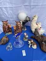 Photo shows mixed animal figurines on blue cloth background including two brown ceramic owls, fluffy llamas, ceramic bird, blue art glass dolphin, clear glass dog, brown dog figure, and small animal figures.