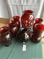 Photo showing the full set of seven ruby red glass vases grouped together on a table, highlighting variety in shapes and sizes.