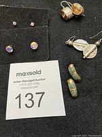Photo showing all five pairs of sterling pierced earrings, including leaf-shaped, goldtone with green stones, embossed oval band style with champagne stones, and two sets of iridescent studs (large and small). Earring styles and stone colors clearly visible.