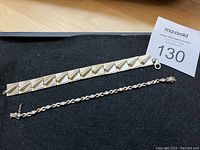 Two silver bracelets laid out flat on black felt with auction lot number tag visible. One bracelet has wide square panels with rolled design, the other has intertwined links with round CZ stones.
