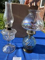 Two vintage oil lamps on a blue surface outside. One lamp is clear pressed glass with a slender chimney and flared base. The other lamp has a blue swirled glass base and an iridescent wide chimney.