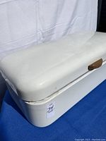 Side angle showing white enameled metal bread box with wood handle on blue cloth backdrop
