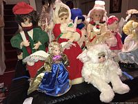 Full view of 5 Christmas dolls arranged together on black cloth, showing dolls in green and red holding electric candles, the small blue dress doll, the white furry doll and the cream/pink dressed doll.