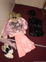 Pink satin 'Pink Lady' jacket, pink skirt, saddle shoes, pink socks, and dark wig on floor with pink carpet background.